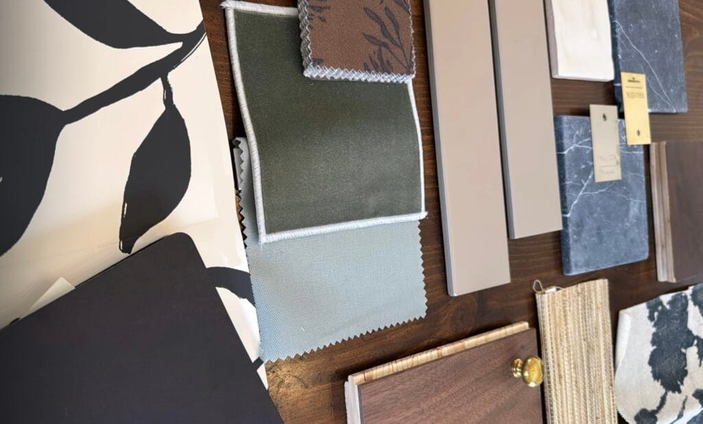 Finding the final touch. A flat lay of interior design material samples arranged on a wooden surface, including olive green and light blue fabric swatches, a small brown patterned fabric, taupe paint or laminate samples, a piece of dark marble, woven beige textile, and a wood panel with a brass knob.