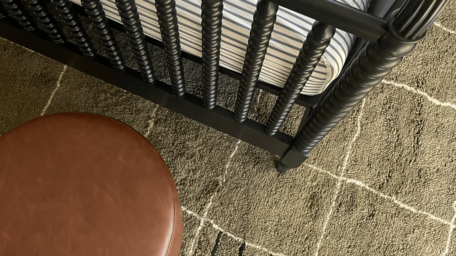 Close-up view of a textured taupe area rug with a subtle grid pattern, a round brown leather ottoman, and part of a black spindle crib with a striped crib sheet.