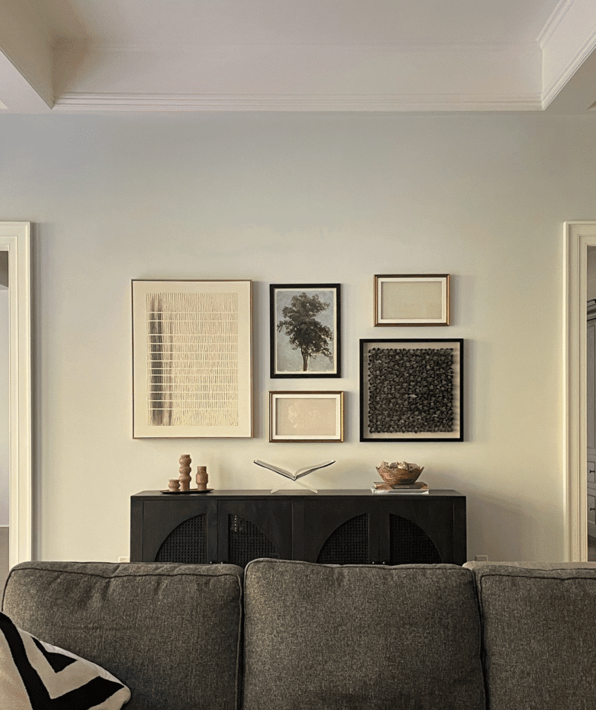 A modern, neutral-toned living room with a gray sofa in the foreground, a black sideboard against a light gray wall, and a curated gallery wall of six framed art pieces above the sideboard. Decorative objects, including stacked wooden pieces, a book on a stand, and a bowl.