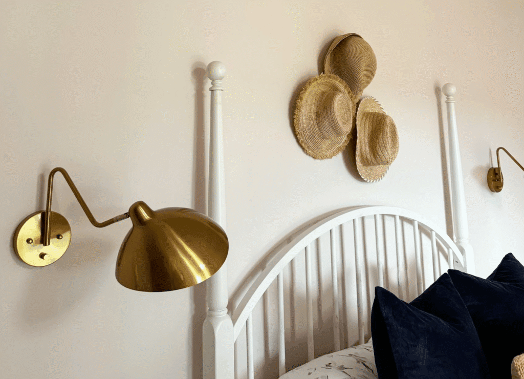 Bedroom designed with soft colors, brass sconce lights, bed, and hats hanging on the wall.